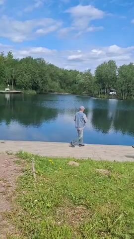 The video shows a man fishing on a sunny day in Kipen, Russia. The scene is set by a calm body of water, likely a lake or large pond, surrounded by lush green trees on the far bank. In the foreground, there is a stretch of green grass, and a wooden deck or pier extends over the water. A wooden bench is placed on the deck to the right.

The man, dressed in a light-colored top (possibly a denim shirt or jacket) and jeans, stands on the wooden deck facing the water. At the beginning of the video, he is seen adjusting or preparing, then he raises a fishing rod and casts his line into the water. He continues to hold the rod, engaged in fishing.

The sky is bright blue with scattered white clouds, suggesting clear and pleasant weather. The water is still, reflecting the sky and the surrounding greenery. A small, light-colored building is visible among the trees on the far side of the water, adding a touch of distant human presence to the otherwise natural landscape. The overall atmosphere appears peaceful and serene.