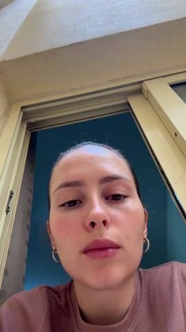 The video shows a woman holding a handheld device to her face. The camera is positioned below her, looking upward. The woman is wearing a brownish-pink top and has her hair pulled back. She is using the device, which appears to be a facial massager or beauty tool with a silver head, near her eye and cheek. Behind her, a doorway or window frame is visible, leading to an outdoor area where pipes run along the ceiling. The lighting is bright, suggesting daylight. The video's perspective and close proximity to the woman focus attention on her face and the beauty device she is using.
