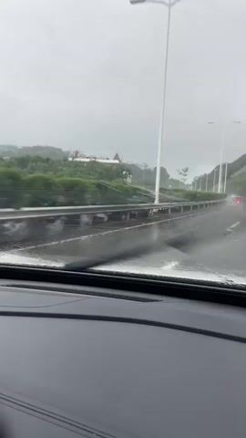 The video shows a view from inside a car driving on a wet road. The weather is overcast, and it appears to be raining, as evidenced by the water droplets on the windshield and the wet surface of the road. The wipers are activated, clearing the windshield intermittently. In the background, there are lush green hills covered in trees, and a few buildings, including what looks like a large, ornate building with red roofs, and some modern high-rise structures. The road has multiple lanes and a metal guardrail separates it from the greenery and buildings on the side. The overall impression is a gloomy, rainy day drive through an urban and natural landscape. No people are visible. The time of day is not clearly indicated, but the overcast sky suggests it could be daytime. There is no visible text in the video.