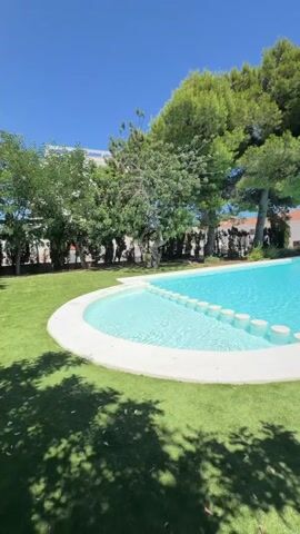 The video pans across a bright, sunny outdoor scene with a large, turquoise swimming pool in the foreground. The pool has a curved edge with white coping and a shallow area featuring a series of white, circular stepping stones. Lush green grass surrounds the pool, with a shadow pattern cast by overhead trees. In the background, there are several white buildings with terracotta roofs, tall green trees, and dense shrubbery. Further in the distance, a person with dark, curly hair wearing a white t-shirt is visible standing in front of some bushes, facing towards a bright blue body of water, presumably the ocean. The sky is a clear, vibrant blue.