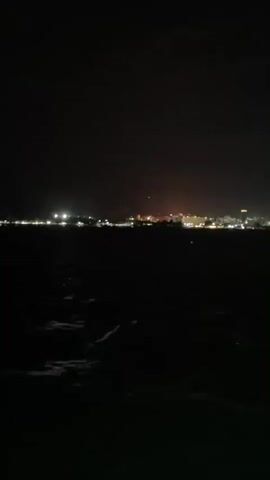 The video shows a blurry view of a city skyline at night. The city is illuminated by numerous lights, mostly white and yellow, with one orange tower in the center. The foreground is almost entirely dark, representing a body of water with some visible waves or ripples. The horizon is obscured, making it difficult to discern specific details of the buildings. The video is shaky, implying it was recorded from a moving position.