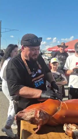 A man wearing a black t-shirt, an apron that says "ХРЕНОВ BBQ", a black beanie, and black gloves is shown in the foreground. He is standing behind a wooden cutting board and appears to be carving a whole roasted pig. The pig is golden brown and positioned on its side. Several people are visible in the background, including a young boy wearing a baseball cap, a woman holding a phone to record, and men wearing hats and t-shirts. The scene appears to be outdoors, possibly at an event or festival, with a bright blue sky and a red canopy visible in the distance. The man is focused on his task of carving the pig.