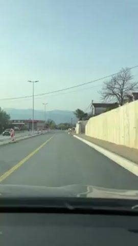 A car is driving down a newly paved road on a clear day. On the left is a building, street lights, and road construction markers. On the right is a wall with a house on top. There are mountains in the distance.