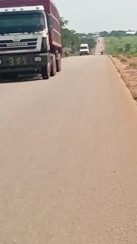 A large truck with a red cargo container is driving down a paved road. The truck has the word MANCHESTER visible on the side of its cab. In the distance, other vehicles can be seen on the road. The surrounding environment consists of sparse vegetation and trees. The sky is partly cloudy. The camera appears to be mounted on a moving vehicle, likely following the truck.