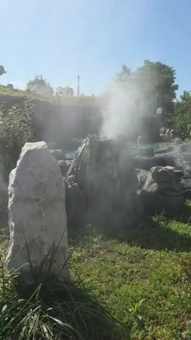 The video shows a steaming rock formation in what appears to be an outdoor park setting. In the foreground, there is a tall, light-colored rock with grass and some small plants around its base. Behind it, a darker, more jagged rock formation is emitting a plume of white steam upwards. To the left of the steaming rock, a body of water, possibly a pool or river with a blue-green hue, is visible, with a building structure featuring a green, grass-covered roof situated behind it. Further to the right, more grey rocks of various sizes are scattered across the landscape, also producing some wisps of steam. The overall environment is green with grass and trees in the background, and the sky is clear and blue. The sun appears to be shining, creating bright lighting conditions.