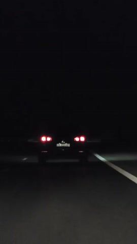 A car drives on a dark road at night. The car's taillights are illuminated, casting red light on the asphalt. The license plate is visible, though the text is unreadable due to motion blur and low light. A white line on the right side of the road indicates a lane marker. The surrounding environment is completely dark, suggesting an open road with no other light sources. The perspective is from behind the car, suggesting it is being filmed from another vehicle following closely.