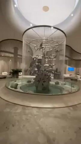 The camera pans across the lobby of a high-end building, showing a central water feature surrounded by a circular glass enclosure. The fountain has abstract metal sculptures resembling fish or marine life. The lobby features smooth, light-colored flooring with some areas in a marble-like pattern. The walls are curved and feature a design with vertical slats behind sheer white curtains. Several white lounge chairs are arranged in the seating area. Plants are strategically placed around the lobby. The ceiling is rounded with recessed lighting. The overall atmosphere is clean, bright, and modern.