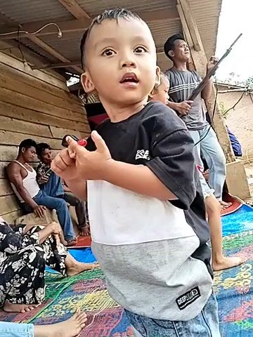 The video shows a young boy in the foreground wearing a black and white t-shirt and jeans. He is holding something red in his right hand and looking upwards. Behind him, a girl in a red shirt and denim shorts is standing, and a man in a dark shirt is behind her holding a rifle. Several other people are visible in the background, some sitting on the ground and others standing. The setting appears to be outdoors, with a wooden structure in the background. The ground is covered with a colorful mat.