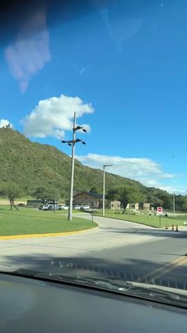 The video shows a view from inside a car driving on a road on a sunny day. The sky is bright blue with scattered white clouds. The car is approaching a section of the road marked by orange traffic cones. To the left, there is a grassy area with a sign that reads "Parque de Encuentro de la Familia". There are buildings and parked cars in the background, with a green mountain further back. As the car continues, it passes under a pedestrian overpass with towers. Other cars can be seen on the road ahead. The overall impression is of a pleasant day for a drive.