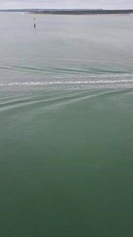 A drone shot shows a boat moving through water. The water is green and has waves behind the boat. A buoy is visible in the distance. The sky is cloudy and grey. The horizon is visible in the background, showing land in the distance.