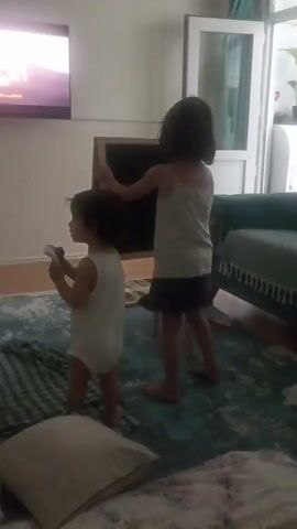 Two young children are in a room. A girl on the left is wearing a white onesie and is holding a toy microphone, looking at a television screen. A taller girl on the right is wearing a white tank top and dark shorts, facing a blackboard on an easel. She appears to be gesturing towards the blackboard. The room has a rug on the floor, a sofa on the right, and a window in the background. The television screen is visible in the upper left corner.