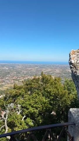 The video captures a panoramic view from a high vantage point, likely a mountain or a tall building, overlooking a sprawling town or city. The sky is a clear, vibrant blue, with no clouds visible. In the distance, the horizon is marked by the faint blue line of the ocean or a large body of water. The town below is characterized by densely packed buildings with terracotta roofs, suggesting an Italian or Mediterranean architectural style. The landscape is hilly and appears to be dotted with trees and fields. In the foreground, there are trees with green leaves, some of which are partially obscuring the view. A dark metal railing runs along the bottom of the frame, indicating the presence of a balcony, terrace, or viewing platform. The camera appears to be static or making very slow, subtle movements, allowing the viewer to take in the expansive scenery. No people or specific actions are discernible in the video. The overall impression is one of a serene and picturesque vista on a sunny day.