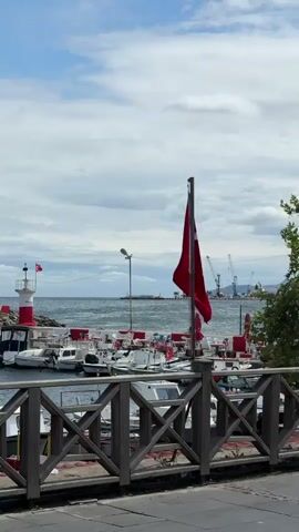 The video captures a view of a harbor with boats docked and a Turkish flag prominently displayed on a flagpole. In the foreground, a wooden railing divides the viewer's perspective from the harbor. A lighthouse is visible in the distance, near the shore. The sky is cloudy, with patches of blue peeking through. An older man, wearing a light blue polo shirt, walks across the frame from left to right in front of the railing and the harbor view. The boats in the harbor are small and appear to be fishing vessels, many adorned with red elements, possibly matching the flag. Further in the background, cranes and industrial structures suggest a larger port or shipyard area. The overall scene conveys a sense of a coastal town or a working harbor.