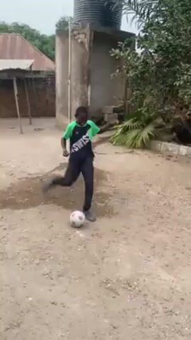 The video starts with a blurred shot of a sandy yard with trees along a brick wall and a blue object. The camera pans to reveal a metal gate between two buildings. A young boy wearing a black and green shirt with a yellow stripe runs into the frame, smiling at the camera. He throws his arms into the air.