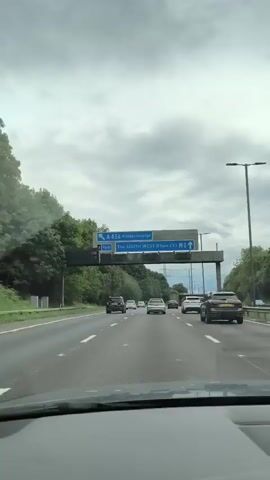 This is a first-person perspective video of driving on a highway. The driver is following a multi-lane highway with other vehicles ahead. Overhead signs indicate directions to A456 Kidderminster and The South West Birmingham (M5). Trees line the left side of the highway, and utility poles are visible on the right. The sky is overcast. The interior of the car, including the dashboard, is partially visible in the foreground.