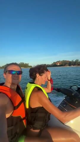 Two people, a man and a woman, are riding a jet ski on the ocean. The man is in the front, wearing sunglasses and an orange life vest. The woman is behind him, wearing a yellow life vest. The water is choppy, and there are trees and buildings on a distant shore. The sky is clear and blue. Another jet ski is visible in the distance.
