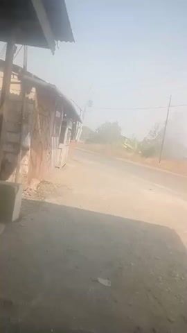 The video shows an outdoor scene with a dirt road and a small, dilapidated building on the left. The building appears to be made of wood and has graffiti on its side. The sky is hazy and bright, suggesting either a sunny day or dusty conditions. The road is unpaved and curves slightly to the right. Trees and other vegetation are visible in the background, partially obscured by the haze. There are power lines overhead. The overall impression is one of a rural or semi-rural environment.