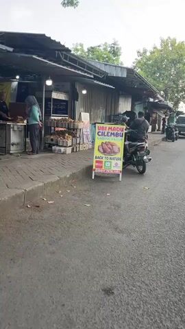 The video shows a street scene with several people and motorcycles. In the foreground, a motorcycle is parked on the side of the road, and a person is sitting on it. A white and green sign is placed on the sidewalk in front of a food stall. The sign has text that reads "UBI MADU CILEMBU" and shows an image of sweet potatoes. Behind the sign, the food stall is visible, with people inside serving food. Further down the street, other motorcycles and people are present, and a small truck is parked on the right side of the road. The environment appears to be an outdoor market or roadside food vendor area, with stalls and sidewalks lining the street.
