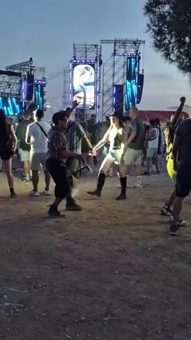 A crowd of people stands in a dirt area in front of a large stage setup with multiple screens and light structures. The sky is dusky, indicating either early morning or late evening. Some individuals are dancing and interacting with each other. The stage is illuminated with blue and green lights, and a large screen displays an image. The overall atmosphere suggests an outdoor event, possibly a concert or festival.