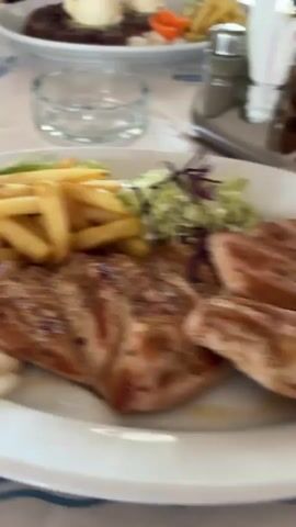 The video depicts a restaurant scene featuring several people enjoying their meals. The camera focuses on dishes containing what appears to be grilled meat, french fries, salad, and small carrots. One plate has what seems to be two scoops of butter on top. Bread is served in a basket on the table. Some people are visible seated, eating their food with forks. The table is covered with a patterned tablecloth, and an ashtray is also present.