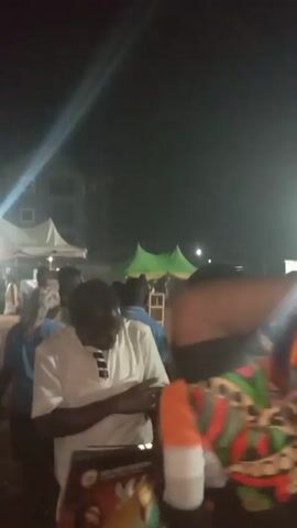 The video, recorded at night, shows a crowd of people gathered under a large, green and yellow tent. There is a bright light in the background. The camera moves somewhat erratically, focusing on the backs of people and occasionally showing more of the crowd. Someone's arm with an orange and purple patterned shirt appears in front of the camera briefly. Music is playing in the background.