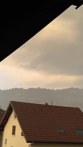 This video captures a view from a balcony or window, looking out towards a mountainous landscape under a cloudy sky. The foreground features the tiled roof of a house with dormer windows and chimneys. In the mid-ground, rolling hills covered in vegetation are visible, with faint lights suggesting buildings or settlements. The background is dominated by the hazy silhouettes of distant mountains, some of which appear to be dusted with snow, indicating a cooler climate or higher altitude.

The sky is overcast with a muted, warm light, suggesting either dawn or dusk. The clouds are heavy and appear to be carrying precipitation, giving the scene a somber or dramatic feel. The overall atmosphere is one of quietude and natural beauty, with the human element present in the form of the house but secondary to the grandeur of the mountains. There is no visible text in the media file. The location context of Cluses, France, places this scene in the French Alps, which aligns with the mountainous terrain and the potential for snow-capped peaks. The weather appears to be overcast, possibly with rain or mist, adding to the moody ambiance.