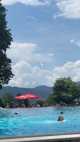A person is swimming in a bright blue swimming pool. In the background, several people are relaxing on lounge chairs under a red umbrella. Behind them, there are green trees and mountains under a clear blue sky with fluffy white clouds. A hang glider is visible in the sky.