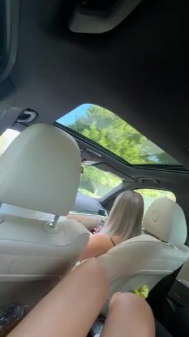 The video, captured from the back seat of a car, shows three young females. The person filming is focusing on their own legs, which are in the foreground. Further back in the car, another female with blonde hair is visible, and a third female with dark hair and glasses is using a smartphone. The car has a sunroof, and through it, green trees and a bright sky are visible, indicating it's daytime and sunny. The interior of the car appears to be made of light-colored leather. The video appears to be a casual, in-car recording.