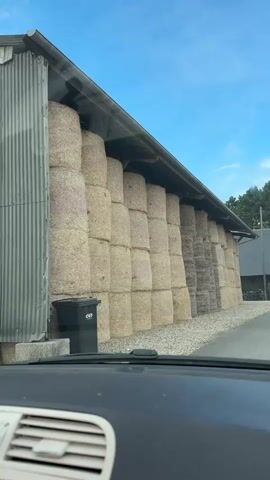 The video shows a side view of a structure with large stacks of hay bales stored under a metal roof overhang. The hay bales are stacked in neat columns. To the left of the hay bales, there is a grey corrugated metal wall and a black trash can. The ground in front of the hay bales is covered with gravel. The video pans to the right, showing more hay bales, some of which appear darker and possibly older than the others. The sky is visible above, with some clouds and trees in the distance. The video is shot from inside a vehicle, as indicated by the dashboard visible in the foreground.