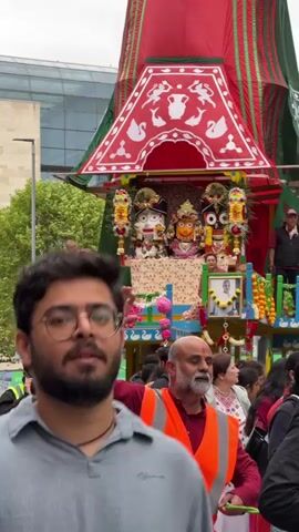A man wearing glasses and a grey shirt is in the foreground, looking towards the camera and speaking. Behind him, a large, decorated chariot is being pulled through a street. The chariot is adorned with red and green fabric, intricate carvings, and images of deities. Several people are visible around the chariot, some wearing bright orange and yellow safety vests. The environment appears to be an urban setting with buildings and trees in the background. The overall scene suggests a religious procession or festival.