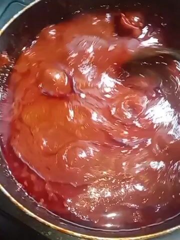 A top-down shot shows a pot of reddish-brown liquid being stirred with a spoon. The liquid is thick and bubbling slightly.  The pot is on a dark surface, likely a stovetop. The spoon is dark and shiny, reflecting light. Small pieces of solid matter are visible within the liquid.