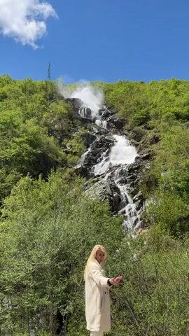 The video shows a woman and a waterfall. The woman, who has long blonde hair and is wearing a light-colored coat, stands in front of a lush green landscape. She appears to be using a smartphone. Behind her, a large waterfall cascades down a rocky hillside covered in dense green vegetation. The sky is blue with a few scattered clouds. A power line tower is visible on top of the mountain.