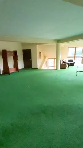The video opens on a wide, dimly lit room with bright green carpeting and a cream-colored ceiling. A staircase is visible on the left, with a dark brown door at its base. In the center of the room, a ping pong table is set up, and further in the background, a foosball table is visible. Two individuals are present, one person is sitting at a table playing a game, while another person is standing near the foosball table. The room features large windows that look out onto a natural, possibly wooded, environment. The overall atmosphere appears to be that of a recreational area.