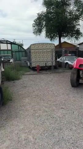 A red Chevrolet Corvette with a lift kit and oversized tires sits parked on a gravel lot. The car is customized to resemble Lightning McQueen from the movie "Cars," with cartoon eyes painted on the windshield and the number 95 on the side. A yellow "CAUTION KEEP OFF" sign is attached to the front bumper. In the background, a tree, a fence, a white vehicle, and buildings are visible. A person is holding a phone, seemingly taking a picture of the car. The sky is overcast.
