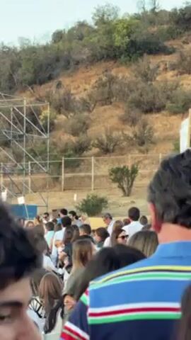The video shows a large crowd of people gathered outdoors at an event. Many of the people are standing or sitting in chairs, facing a stage that is set up with lights and speakers. The stage appears to be made of wooden structures, and there is a screen to the right. In the background is a hillside covered with dry grass and bushes. The sky is bright, suggesting it is daytime. The people in the crowd are wearing a variety of clothing. Some are dressed in casual attire, while others are wearing more formal outfits. The camera is panning from left to right.