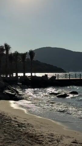 This media file captures a serene coastal scene at Bodrum, Turkey. The primary subjects are a sandy beach in the foreground, with gentle waves lapping at the shore, and a series of floating cabins or bungalows situated on the water in the middle ground. These structures are dark in color, with visible windows and ladders extending into the water, suggesting they are accommodations for visitors. A wooden pier connects these cabins to the shore, adorned with a rope railing. In the background, a silhouetted mountain range forms a dramatic backdrop against a clear, bright sky.

The activity taking place is the natural movement of the sea, with waves creating a rhythmic motion and foam. The lighting suggests it is daytime, likely late morning or early afternoon, given the bright sunlight reflecting off the water and the clear, shadow-casting sun. The overall mood is peaceful and tranquil, evoking a sense of relaxation and natural beauty. The presence of the floating cabins hints at a resort or a unique accommodation experience. There is no visible text in the media.