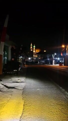 The video shows a nighttime view of a street with a mosque in the background. The mosque is illuminated with lights, highlighting its dome and minarets. There are flags on poles along the street, and a car is parked on the left side. The road is paved, and there are streetlights visible. The overall scene depicts a quiet street at night, with the mosque serving as a prominent landmark.