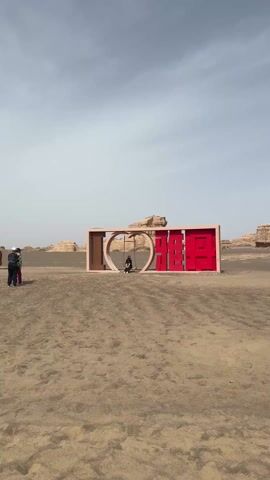 The video shows a desert landscape with people exploring the area. The scene features a large red and tan structure with a person sitting on a swing inside a heart-shaped opening. The camera pans to reveal more of the desert environment, showing individuals interacting with different installations and taking photos. A small wooden hut is also visible in the background. People are walking along a paved path near the hut, observing the landscape.