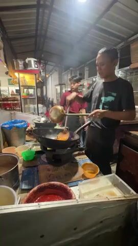 Two young men are preparing food at an outdoor food stall at night. The man in the foreground, wearing a dark t-shirt and apron, is actively cooking in a large wok placed on a gas burner. He is stirring the food with a ladle. The man in the background, wearing a maroon t-shirt, is also involved in food preparation, holding a ladle over another wok. The stall is set up with various containers of ingredients and condiments, including a large bowl of red sauce in the foreground. The environment appears to be a busy street food area with overhead lighting.