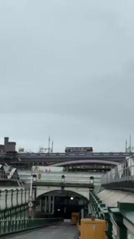 The video shows a view in Edinburgh, Scotland on a cloudy day. In the background is a combination of railway structures, including bridges, arches, and what appears to be Waverley Station. Edinburgh Castle is visible on a hill behind the railway. The foreground shows a section of a bridge or elevated walkway with green and white railings, including a light post with a decorative lantern. A sign with a speed limit of 5 is visible. There is no activity or people apparent in the video.