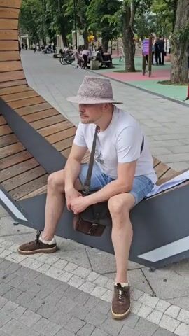 The video shows a man sitting on a modern, curved wooden bench in a park. He is wearing a white t-shirt, blue denim shorts, a light-colored fedora hat, and brown sneakers. A brown messenger bag is slung across his body. The man is looking off to his left, with his hands clasped in his lap. He appears calm and relaxed.

The setting is an outdoor park area with paved walkways and mature trees. In the background, other people can be seen walking and sitting, some on benches. There are also elements of playground equipment and colorful railings visible in the distance, suggesting a recreational space. The lighting indicates it is daytime, likely late morning or afternoon, with diffused sunlight filtering through the trees. The weather appears to be pleasant, suitable for outdoor activities.

The paved ground in front of the bench is a patterned grey and white tile. A large sheet of white paper is visible beneath the man on the bench, potentially a document or a drawing. The overall scene conveys a sense of leisure and public space utilization.