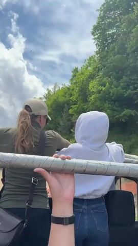 The video begins with a first-person perspective shot looking over the shoulders of two individuals sitting in what appears to be an open-top vehicle. They are facing away from the camera, looking out at a lush green, tree-covered mountainside under a cloudy sky. The person in the foreground is wearing a khaki baseball cap and a green shirt, with their hair tied back. The person behind them is wearing a white hooded sweatshirt and blue jeans. The camera is positioned to capture a metal bar in the foreground, which one of the individuals is holding onto. The scene transitions to a view from the back of the vehicle, showing the road ahead. The vehicle is on a dirt road winding through a mountainous, wooded area. Another similar vehicle is visible ahead, kicking up dust as it drives along the road. The sky is partly cloudy, and the overall environment is natural and rural.