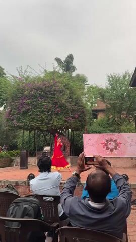 In a medium-shot video, a woman in a red and gold traditional dress dances on a brick stage. The stage is outdoors, with a large tree covered in purple flowers behind it, and a banner to the right of the dancer. Several people sit in black chairs in front of the stage, watching the performance. Two people in the foreground hold up their phones to record the dancer. The sky is overcast. There is a decorative bronze sculpture of a seated figure with multiple arms on the left of the stage.