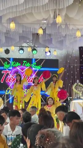 The video captures an indoor wedding reception in Bình An, Vietnam. Four young women, dressed in vibrant yellow áo dài-style outfits, are performing on a stage. Each performer holds either a bouquet of yellow or pink flowers. Behind them, a large LED screen displays the Vietnamese text "Sự Kiện Tiệc Cưới" (Wedding Event) prominently, followed by "Tiên Danh" and a phone number "03348679". To the right of the performers, a man is visible playing a keyboard, providing musical accompaniment.

The event hall is decorated with white draped fabric on the ceiling, giving a soft, cloud-like effect, interspersed with hanging yellow lamps. Stage lighting, including spotlights, illuminates the performers. To the left of the stage, a multi-tiered wedding cake is visible on a table. To the right, another decorative structure, possibly a champagne tower or gift display, stands. Numerous guests are seated at tables in the foreground, facing the stage and watching the performance. The overall atmosphere appears festive and celebratory.