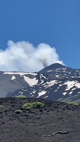 The video shows a volcanic landscape under a bright blue sky with white clouds. In the foreground, there is a dark, rocky terrain with patches of green vegetation. In the background, a large mountain peak is visible, partially covered in snow. A plume of dark smoke is rising from the summit of the mountain, indicating volcanic activity. The smoke billows upwards and mixes with the white clouds. The overall scene depicts a dynamic and natural environment with a volcano actively releasing smoke.