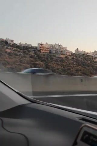The video shows a view from inside a car looking out at a highway. On the right side of the road, there is a concrete barrier. Beyond the barrier, a hillside is covered with scrubby vegetation and dotted with white buildings. The sky is a pale, hazy blue. Several cars are driving on the highway, including a gray SUV in the foreground and a white car in the distance. The video captures a brief moment of travel, with the landscape passing by.