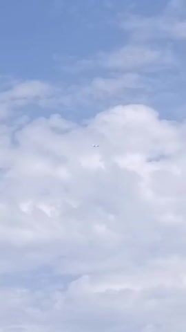A low-angle shot of a bright blue sky with scattered white clouds. A small, distant airplane is visible in the center of the frame, appearing to fly horizontally across the sky. The plane is white and its details are indistinguishable due to the distance. The clouds are fluffy and have a slight texture, suggesting they are cumulus clouds. The overall lighting is bright, indicating it is daytime.