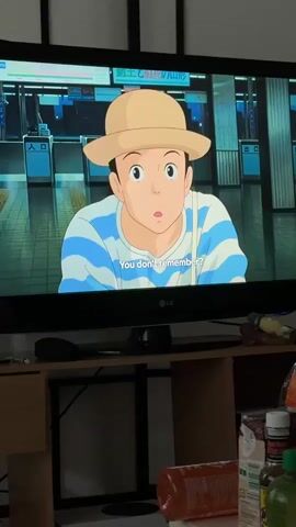 A video shows a television screen displaying an animated scene from what appears to be a Japanese anime or film. Two characters are visible: a female character with a straw hat and a blue and white striped shirt, and a male character in a red cap and a pink hoodie. They are positioned in what looks like a train station or subway entrance, indicated by turnstiles and signs with Japanese text, such as "ステーション デパート" (Station Department Store) and "改札" (ticket gate). English subtitles read, "Of course you don't." In the first frame, the female character is looking forward with a surprised expression. In the subsequent frames, the male character is shown interacting with the female, at one point bowing his head. The television is an LG brand, and it is placed on a wooden TV stand. Below and around the TV, various household items are visible on the stand, including several bottles (one orange plastic bottle, other smaller bottles with labels), and a charging cable. The indoor lighting suggests it is daytime.