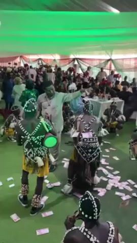 The scene appears to be at an event under a large white tent with green lighting. A crowd of people is gathered, mostly in the background. In the foreground, several performers wearing traditional tribal costumes with beaded decorations and drumming instruments are dancing. A man in a light-colored outfit is among them, directing or interacting with the dancers while throwing money on the floor. The floor is covered with scattered banknotes.