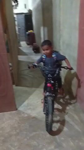 In this video, a young boy wearing a backpack is practicing riding a bicycle indoors. The bicycle is black and red with training wheels. The boy is wearing a dark blue t-shirt and black pants. He appears to be concentrating on riding the bike.

The setting seems to be a room with concrete floors and walls, possibly a hallway or an indoor play area. There are doors and furniture visible in the background, and a pink wall on the right side of the frame. The lighting suggests it might be daytime, but indoors. The overall mood appears to be one of a child learning a new skill, with a sense of focus and determination.

There is no visible text in the video.